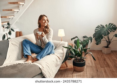 Young woman with a cup of hot coffee or tea sitting on a sofa in a cozy living room at home relaxing and unwinding on a weekend day. - Powered by Shutterstock