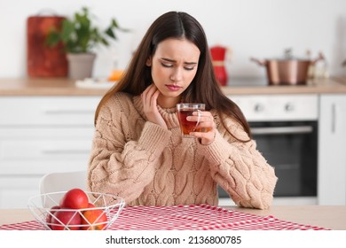 Young Woman With Cup Of Tea Suffering From Sore Throat At Table In Kitchen