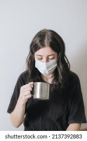 Young Woman With A Cup Of Tea At The Window During Quarantine Wearing Face Mask Worried About The New Lifestyle. Anxiety, Stress, Mental Health, Crisis, Depression, Psychology Therapy.