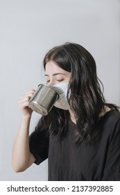 Young Woman With A Cup Of Tea At The Window During Quarantine Wearing Face Mask Worried About The New Lifestyle. Anxiety, Stress, Mental Health, Crisis, Depression, Psychology Therapy.