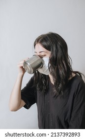 Young Woman With A Cup Of Tea At The Window During Quarantine Wearing Face Mask Worried About The New Lifestyle. Anxiety, Stress, Mental Health, Crisis, Depression, Psychology Therapy.