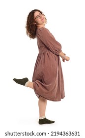 Young Woman With Curly Hair In A Dress, In Glasses Standing Sideways Lifting One Leg Up.  On A White Background