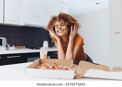 Young woman with curly hair enjoying a delicious homemade pizza while sitting on the kitchen counter - Powered by Shutterstock