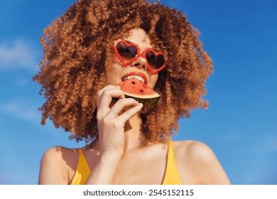 Young woman with curly hair enjoying watermelon on a sunny beach with a clear blue sky Her playful heart shaped sunglasses add a fun touch - Powered by Shutterstock