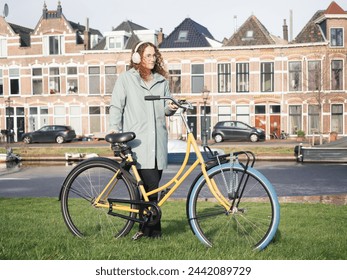 A young woman with curly hair and headphones stands by her colorful bicycle in front of a picturesque row of houses, reflecting a relaxed urban lifestyle on a clear sunny day - Powered by Shutterstock