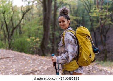 A young woman with curly hair in her 20s exploring the forest trail during autumn, wearing warm clothing and equipped with a yellow backpack, trekking poles, and binoculars.