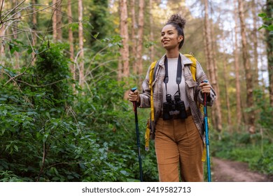 A young woman with curly hair in her 20s exploring the forest trail during autumn, wearing warm clothing and equipped with a yellow backpack, trekking poles, and binoculars. - Powered by Shutterstock