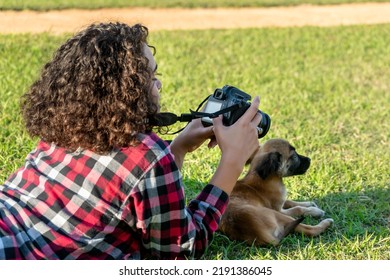 Young Woman With Curly Hair Holding A Digital Camera Within Her Hands, She Is Looking At The Screen, And Lying On The Grass On Her Belly, Elbows On The Groun, A Small Dog Is Lying Down In Front Of Her