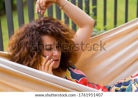 young woman with curly hair is lying in a hammock and talking on a smartphone.