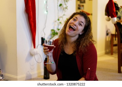 Young Woman With Curly Hair Making A Goofy Face Inside Home During Christmas Holding A Glass Of Sparkling Red Wine And Wearing A Red Sweater.