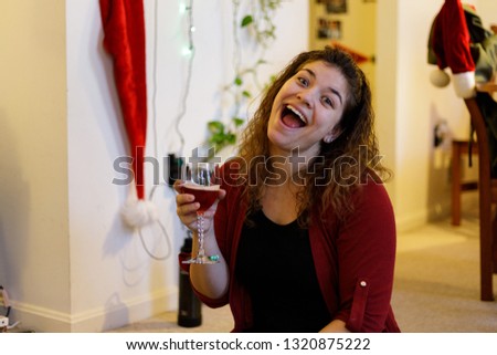 Young woman with curly hair making a goofy face inside home during Christmas holding a glass of sparkling red wine and wearing a red sweater.