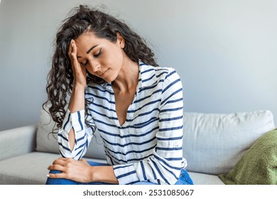 A young woman with curly hair is sitting on a sofa, holding her head in stress. She appears worried and thoughtful, wearing a striped shirt in a calm home setting. - Powered by Shutterstock