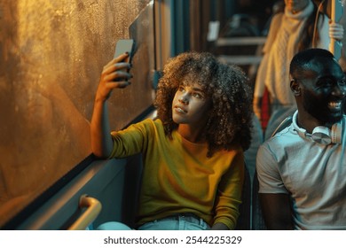 Young woman with curly hair sitting next to a young black man is taking a selfie with her phone on a bus at night. - Powered by Shutterstock