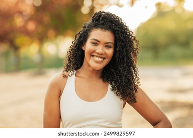 A young woman with curly hair smiles confidently while standing in a sunlit park. The background features trees and a warm, inviting atmosphere typical of the late afternoon. - Powered by Shutterstock