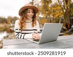 Young woman with curly hair using laptop in the autumn park. Portrait of a young freelance women working outdoors. E-learning concept. Remote work.