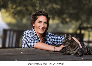 Young woman with curly hair, wearing a checkered shirt, smiles while petting a tabby cat lying on a wooden table in a park-like setting, with trees in the background - Powered by Shutterstock