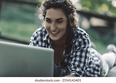 Young woman with curly hair, wearing a checkered shirt, lies on her stomach outdoors, smiling as she works on her laptop, possibly enjoying the fresh air and natural light while staying connected - Powered by Shutterstock