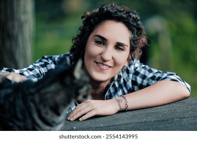 Young woman with curly hair, wearing a checkered shirt, smiles at the camera while petting a tabby cat outdoors, resting on a wooden surface - Powered by Shutterstock