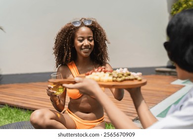 Young woman with curly hair wearing orange bikini holding a drink, while her friend offers her a tray of snacks by the poolside - Powered by Shutterstock