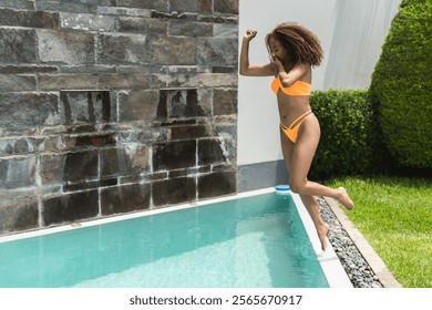 Young woman with curly hair wearing an orange bikini jumping into a turquoise swimming pool on a sunny day - Powered by Shutterstock