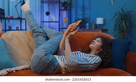 Young woman with curly hairstyle lying on sofa uses mobile phone smiles at night evening home. Girl texting share messages content on smartphone social media applications online watching relax movie - Powered by Shutterstock