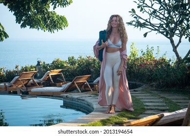 Young woman with curly long blonde hair and blue eyes in white leggings walking with yoga mat in hands on edge of swimming pool on cliff with ocean view in Bali - Powered by Shutterstock