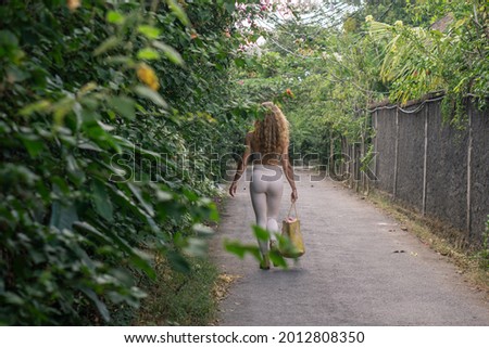 Young woman with curly long blonde hair walking on street with pink flowers in white leggings and small top with beach bag in Bali