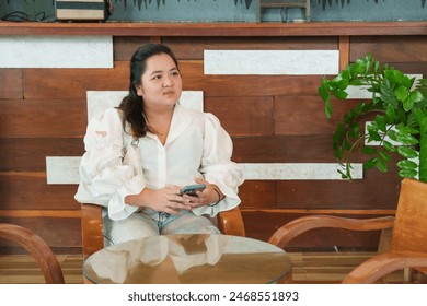 Young woman customer with smartphone, seated, contemplative in cozy coffee shop environment, wooden decor framing natural ambience. small family business - Powered by Shutterstock