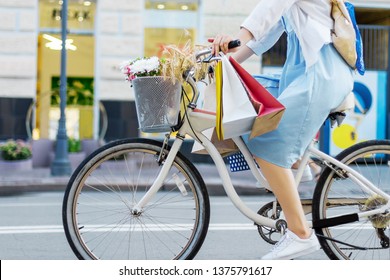 Young Woman Is Cycling Around City On White Bicycle. Girl In Blue Dress Is Going Shopping With Colorful Packages On Handlebars Of Bike. Female Retro Bicycle With Basket With Bouquet Of Flowers.