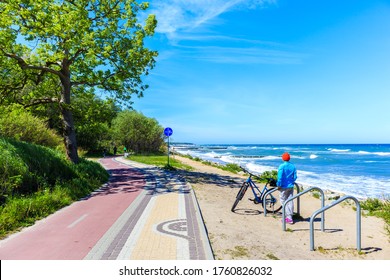 Young Woman Cyclist With Bike Standing Near Cycling Way And Looking At Beautiful White Sand Beach With Blue Sea Near Kolobrzeg Town, Baltic Sea Coast, Poland