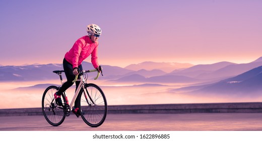 Young Woman Cyclist Riding Road Bike On The Road In The Beautiful Mountains At Purple Sunset. Adventure, Travel, Healthy Lifestyle And Sport Concept.