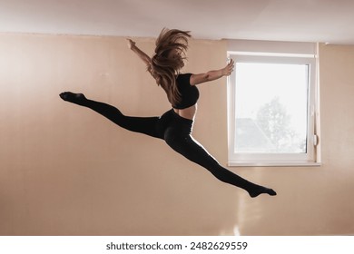 A young woman dancer in black clothing is performs a powerful split leap through the air during a dance rehearsal in a studio. - Powered by Shutterstock