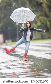 Young woman dances in the rain in the park, holding an umbrella, wearing rainboots.