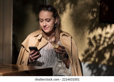 Young Woman With Dark Blonde Hair Smiling And Sitting Outside In Her Balcony Looking At Her Phone And Reading Some Good News While Drinking A Glass Of White Wine, Me Time And Relaxation