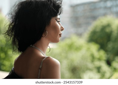 Young woman with dark, curly hair and silver hoop earrings stands on a balcony, gazing contemplatively at the blurred greenery and urban buildings in the background - Powered by Shutterstock