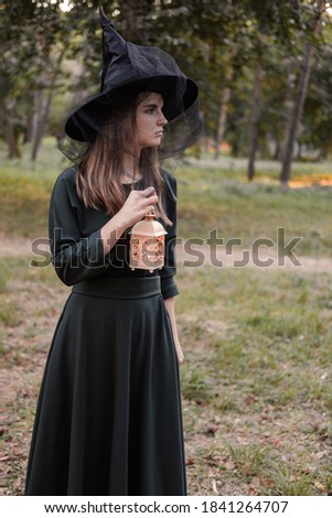 Young woman in dark dress and witch's hat holds lantern with candles in her hands and illuminates the forest. Halloween party costume. Park with autumn trees.