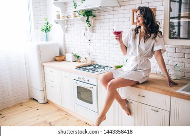 Young Woman With Dark Hair Sitting In Kitchen And Drinking Red Wine From Glass. Enjoying Moment. Alone In Room. Posing In Morning Gown.