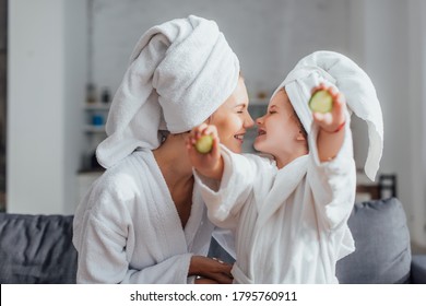 young woman with daughter in white bathrobes and towels on heads sitting face to face while girl holding fresh cucumber slices - Powered by Shutterstock