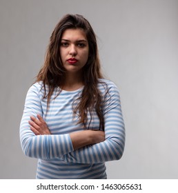 Young Woman With A Deadpan Stern Expression And Penetrating Stare Standing With Folded Arms Looking At The Camera Over A Grey Studio Background
