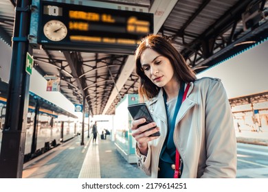Young woman with departure times behind her waiting for her train while holding her mobile phone - Woman looking at the clock in the train station while her train is delayed - Transportation concept