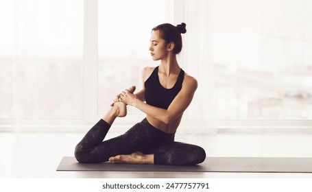 A young woman is depicted sitting on a yoga mat in a well-lit studio. She is dressed in black athletic wear and is performing a yoga pose, maintaining a focused yet serene expression