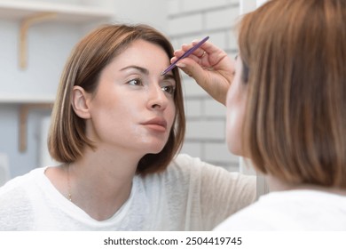 Young woman depilates her eyebrows and gives them the right shape with eyebrow tweezers in front of the mirror. Beauty and self care concept - Powered by Shutterstock