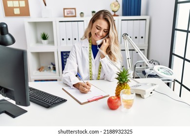 Young Woman Dietician Talking On Telephone Writing On Document At Clinic