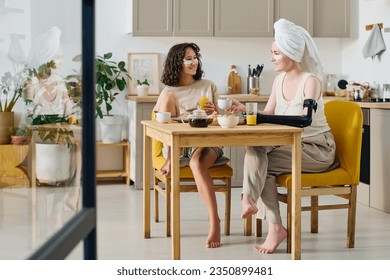 Young woman with disability and her girlfriend with under eye patches sitting by kitchen table, chatting and having coffee and orange juice - Powered by Shutterstock