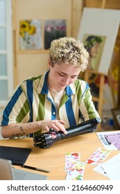 Young Woman With Disability Sticking Handmade Stickers On Her Myoelectric Arm While Sitting By Table In Living Room