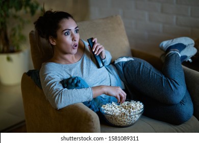 Young woman in disbelief watching TV while eating popcorn in the evening at home. 
