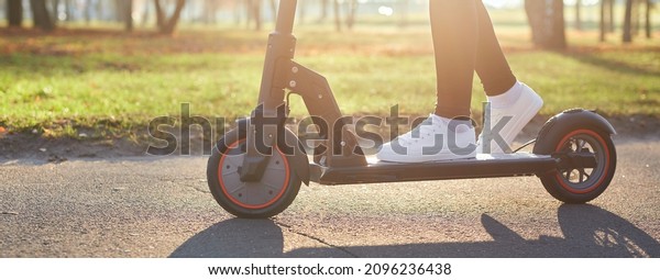 Young woman discover city and park at sunset
with electric scooter or e-scooter. Female Legs in sports sneakers
stand on electric scooter. Girl riding on Ecological and urban
transport.