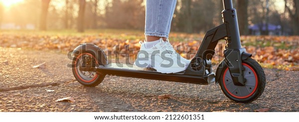 Young woman discover city and park at sunset
with electric scooter or e-scooter. Female Legs in sports sneakers
stand on electric scooter. Girl riding on Ecological and urban
transport.