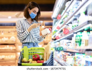 Young Woman In Disposable Face Mask Taking Dairy Products From Shelf In The Supermarket, Holding Bottle And Smartphone, Scanning Bar Code On Product Through Mobile Phone, Walking With Trolley Cart