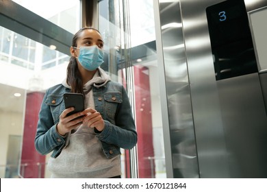 Young Woman With Disposable Mask And Smartphone In Elevator. Dangerous Virus